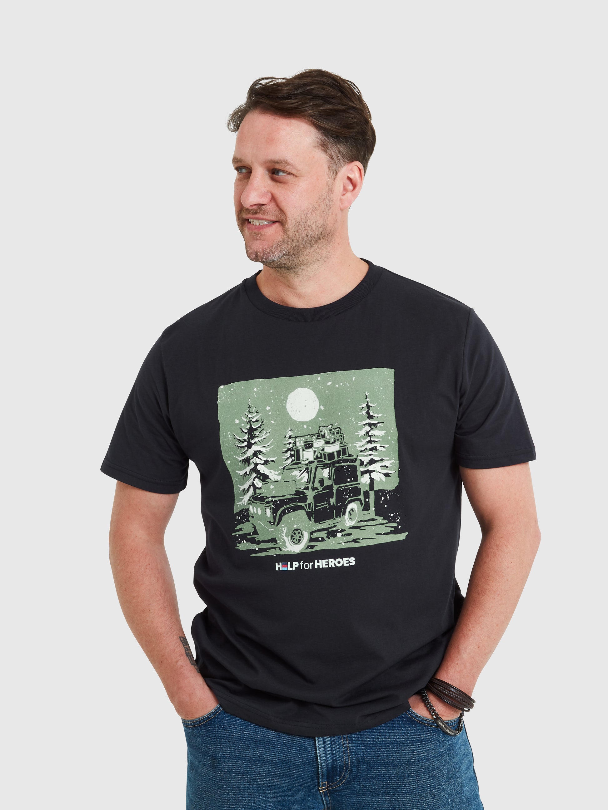 Model wearing a navy Help for Heroes tshirt, featuring a Land Rover carrying Xmas presents through Christmas trees and a snowy scene