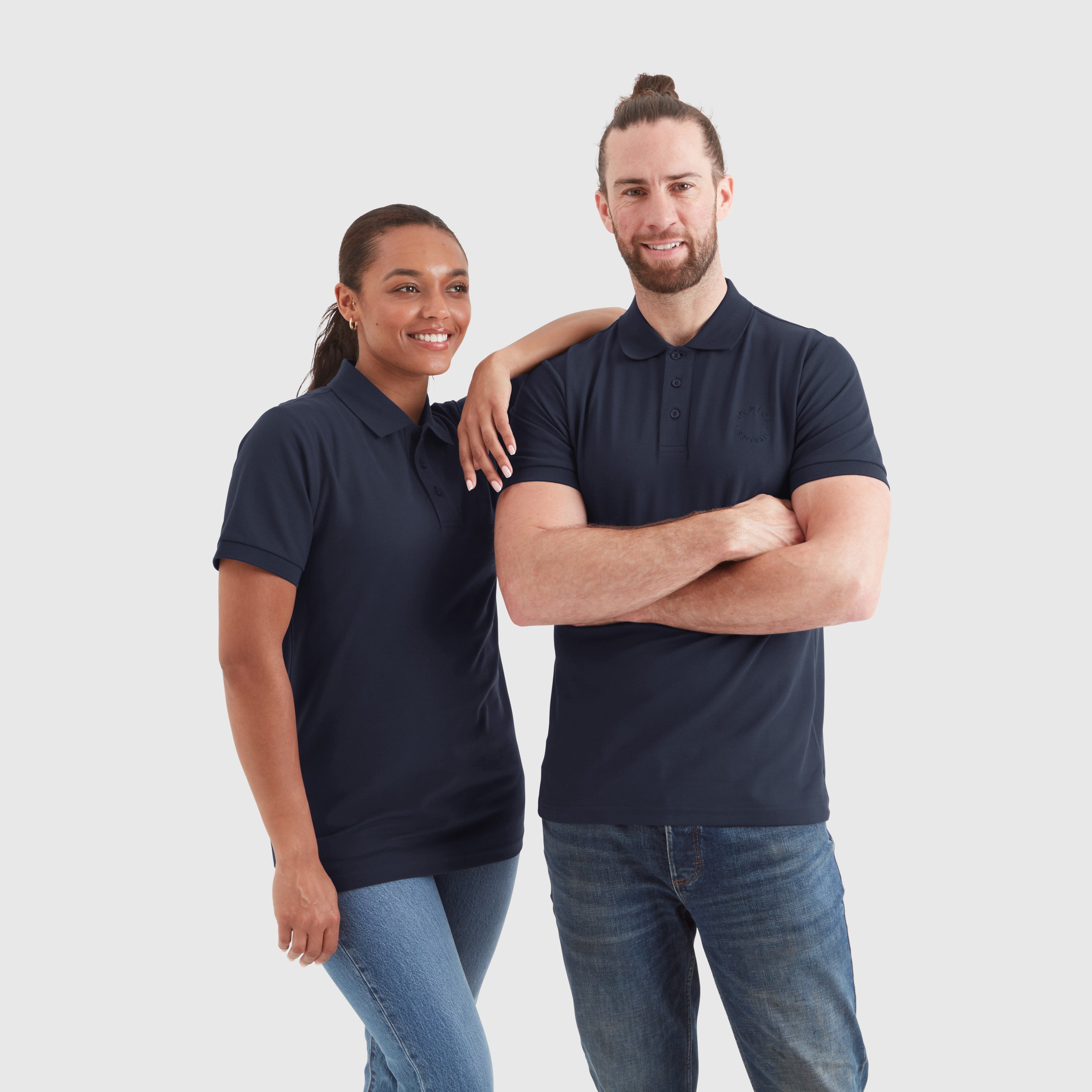 A male and a female model wearing navy pique polo shirts with a subtle circular Help for Heroes logo on the chest and a rubber Heroes stack patch at the back neck.