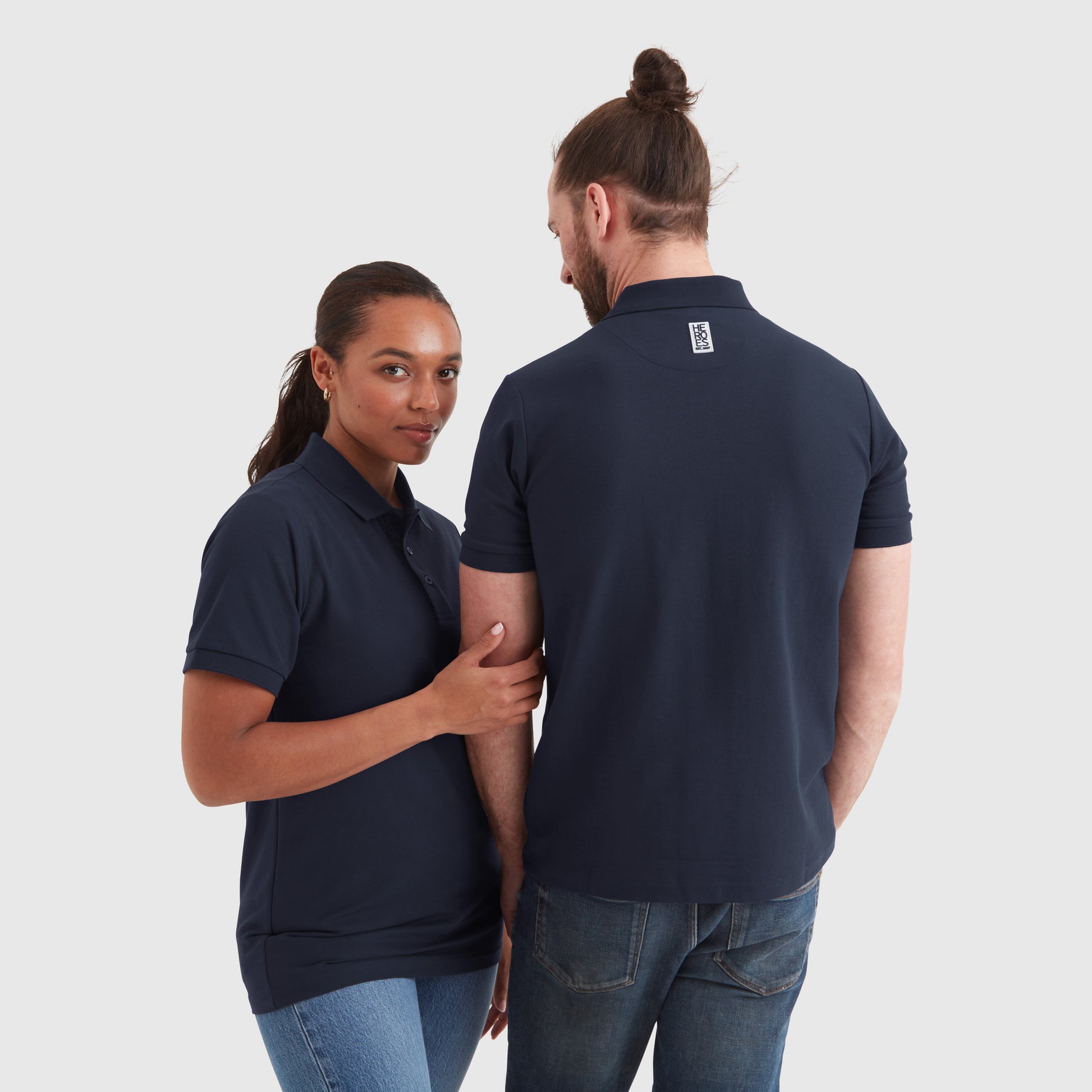 A male and a female model wearing navy pique polo shirts with a subtle circular Help for Heroes logo on the chest and a rubber Heroes stack patch at the back neck.
