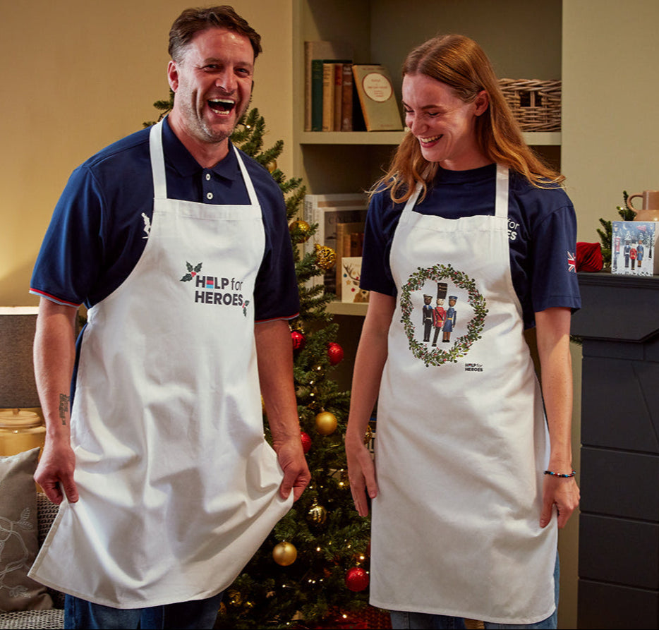 Two models wearing Help for Heroes Christmas aprons, featuring military figures in a wreath design, and Holly Help for Heroes logo design