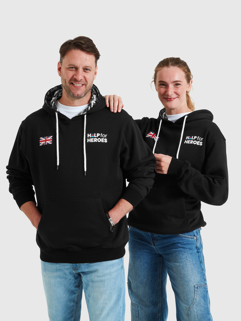 Models wearing black pullover hoodies which feature printed Help for Heroes logo and union Jack flag, and camo detail hood 