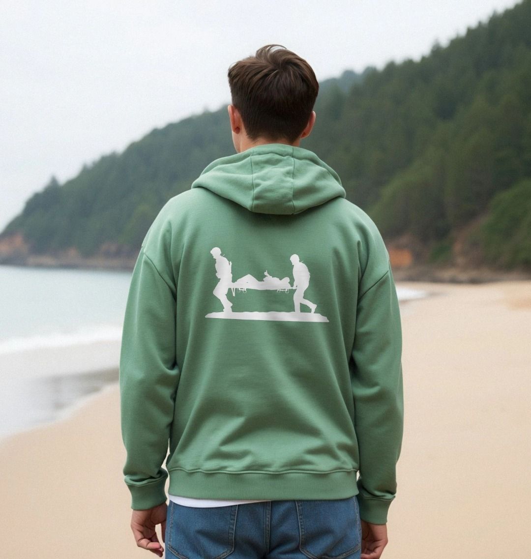 Person wearing a surf green hoodie with a graphic on the back, standing on a beach with a forested area in the background.